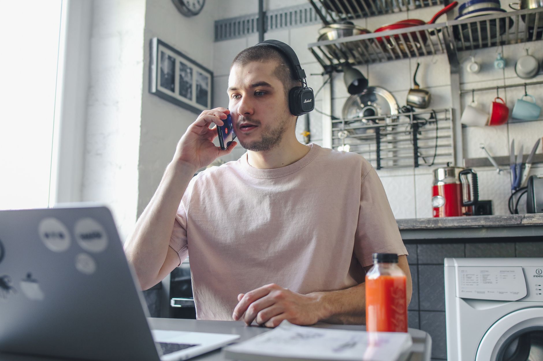 Un uomo seduto a un tavolo in cucina, con cuffie e telefono all'orecchio, davanti a un computer portatile aperto