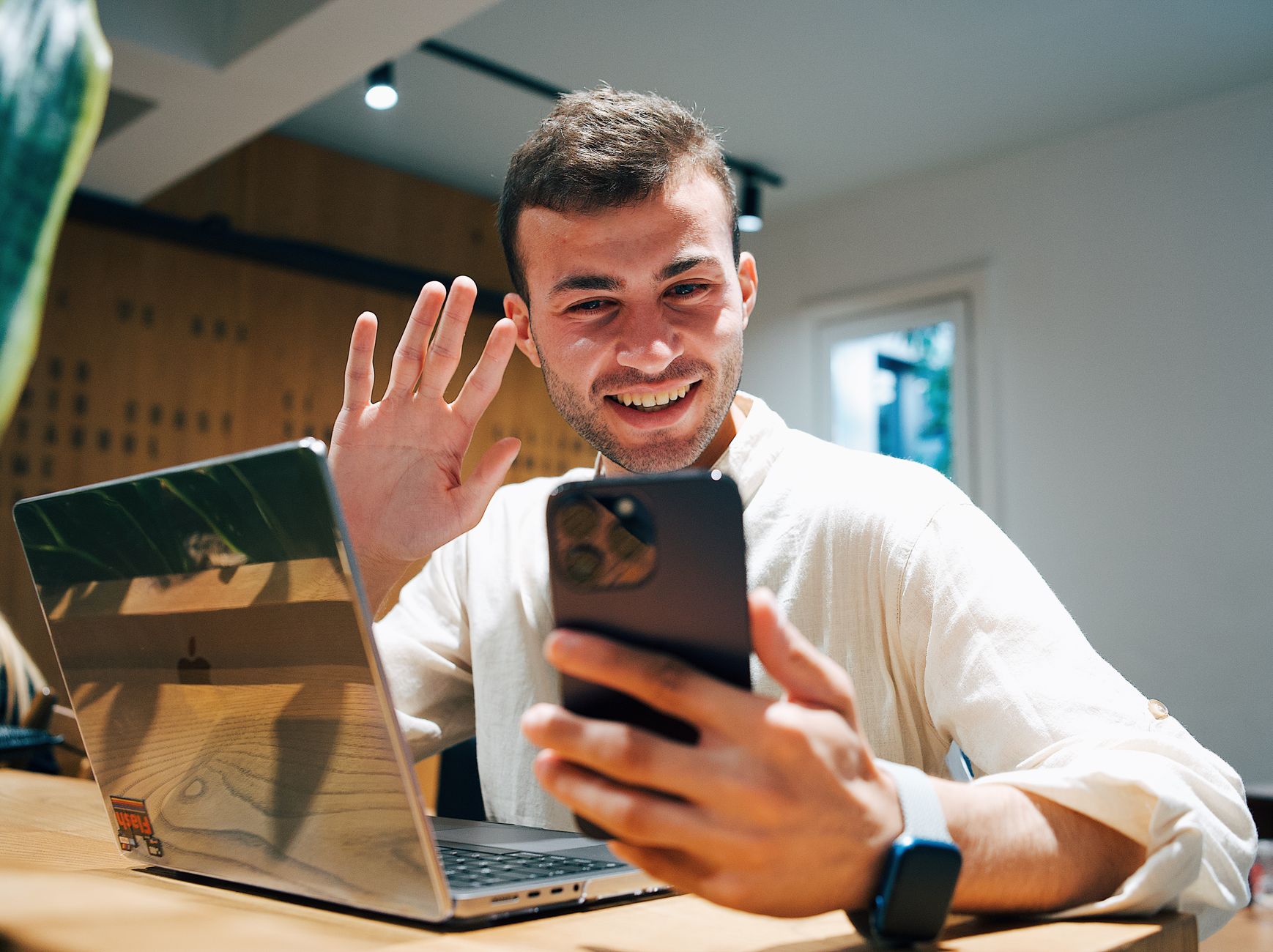 Un giovane sorridente saluta con la mano durante una videochiamata sul telefono, con un portatile accanto